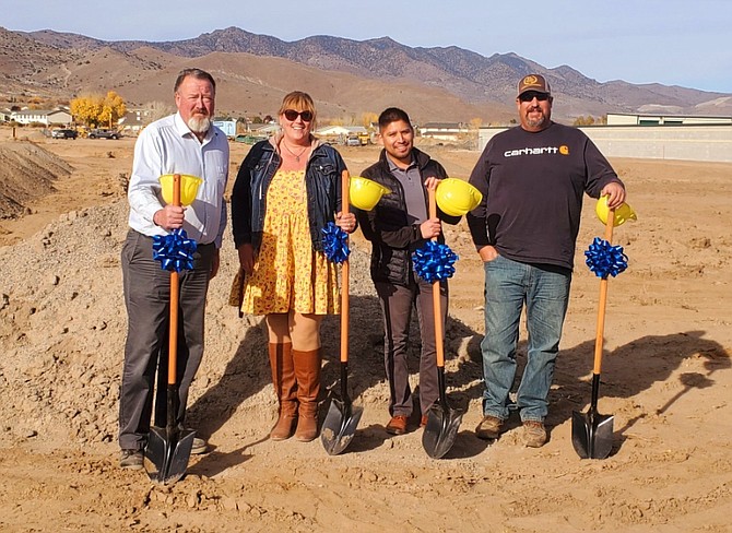 Four people holding ceremonial shovels with ribbons, standing on a construction site with a mountain range in the background.